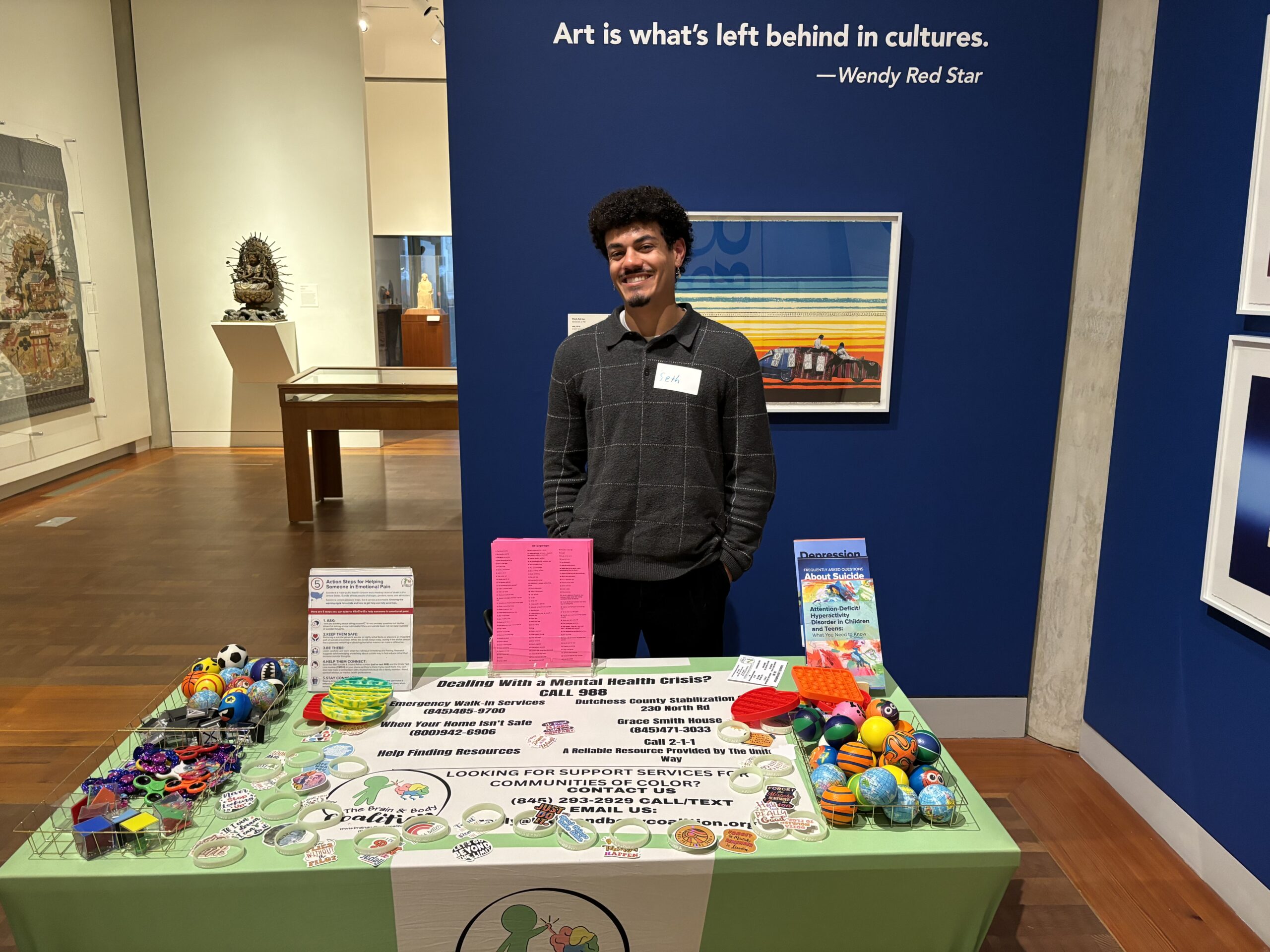 A YOUNG MAN STANDS IN FRONT OF A TABLE SHOWCASING VARIOUS ITEMS